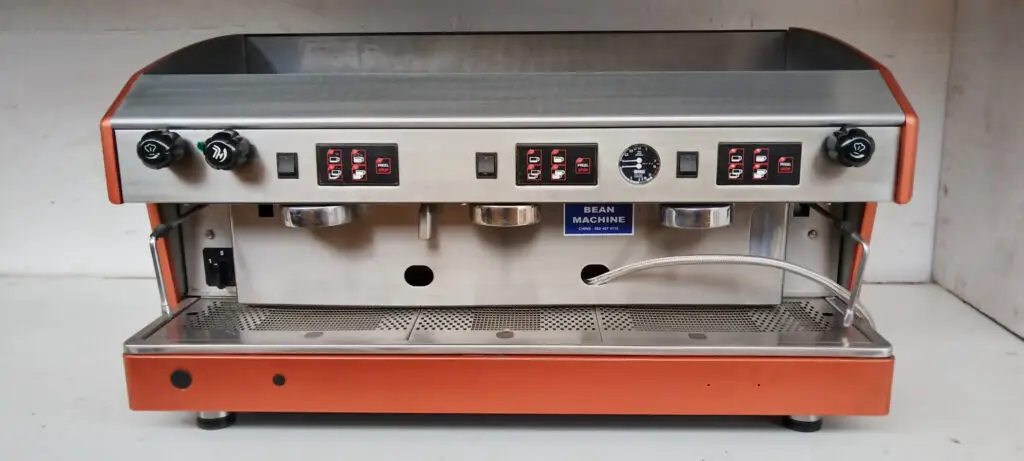 Industrial espresso machine with stainless steel body and orange sides, featuring multiple brew group heads and control panels on the front.