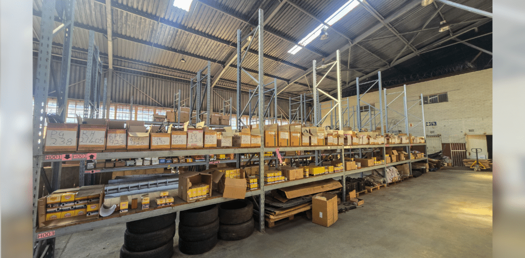 Industrial warehouse aisle with tall metal shelving, labeled boxes, and tires on the floor beneath the lower shelf; bright overhead lighting and concrete floor.