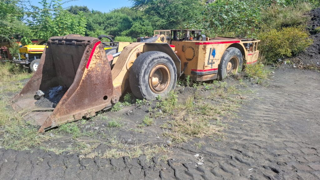 Old, rusted front-end loader with a large bucket resting on a muddy, weed-filled lot; greenery and a yellow vehicle visible in the background.
