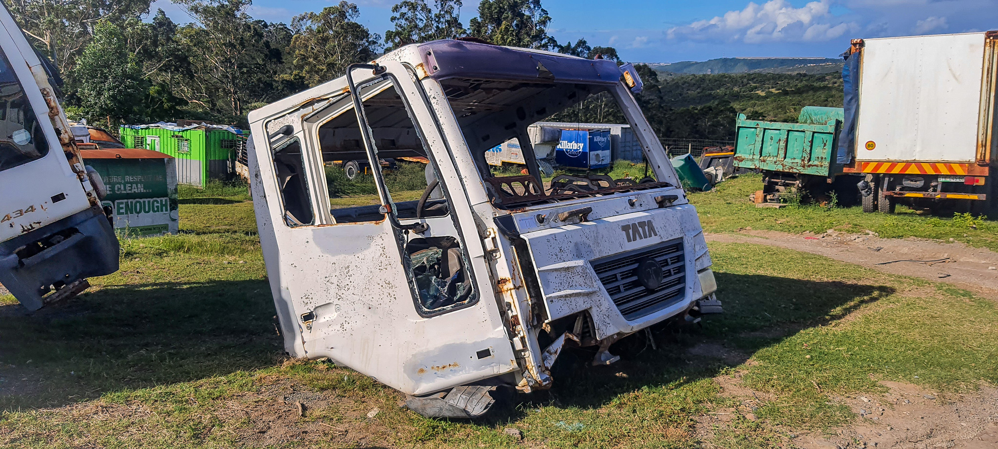 White Tata truck cabin tipped on its side in a junkyard, rusted and weathered, with other scrap trucks and green storage containers in the background