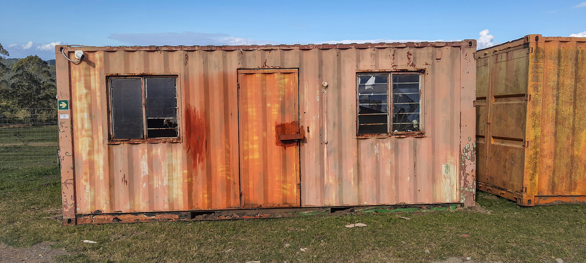 Rusty orange shipping container building with two windows and a central door on a grassy field under a blue sky.