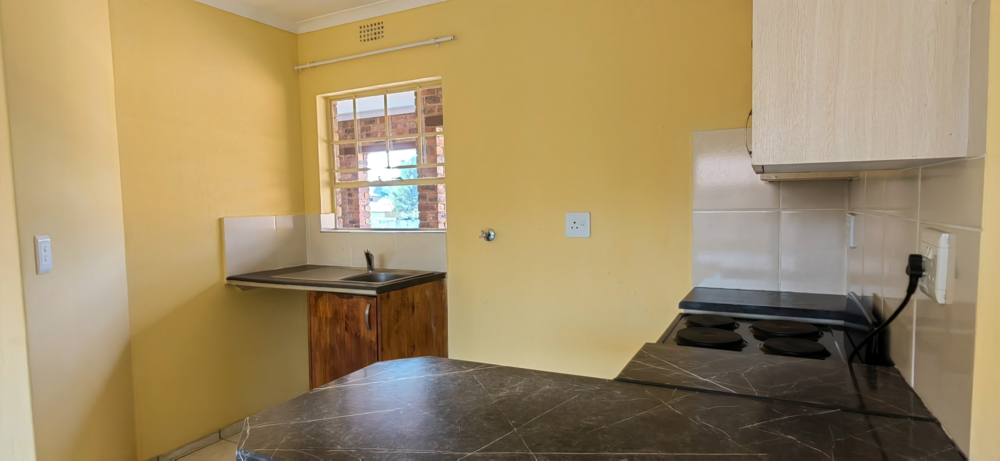 Small kitchen with yellow walls, window letting in light, sink, and wooden cabinet under the counter with a stovetop to the right.