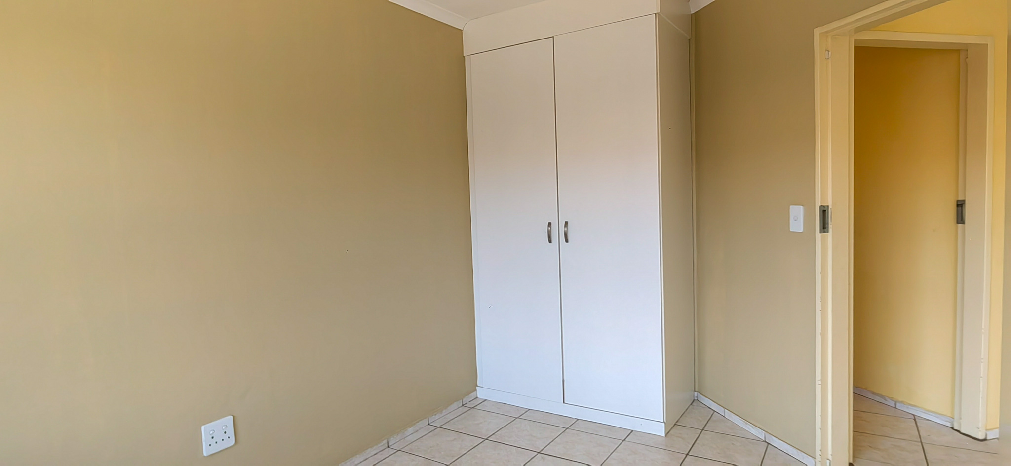 Tall white double-door wardrobe against a beige wall in a tiled room corner, with a doorway on the right.